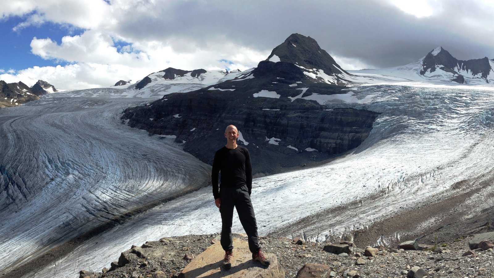 Jed Anderson standing on a rock outcrop in front of a glacier in the Canadian Rockies, with snow-covered mountain peaks in the background.